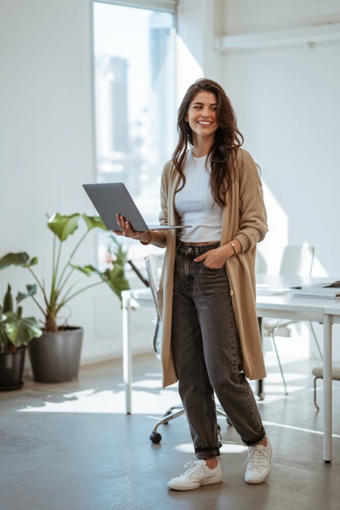 Trendy girl in dark-wash straight-leg jeans, long beige cardigan, tucked-in top, and casual tennis shoes, smiling in a relaxed office