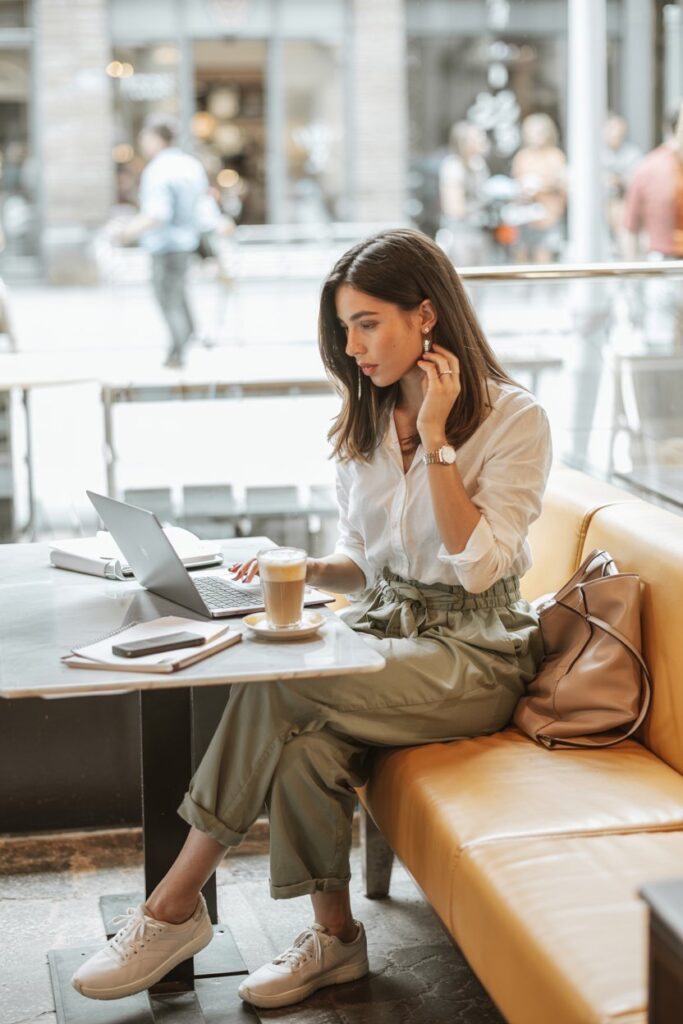 Trendy girl in paperbag waist pants, a tucked-in white button-up shirt, and neutral tennis shoes, seated at a coffee shop with her laptop