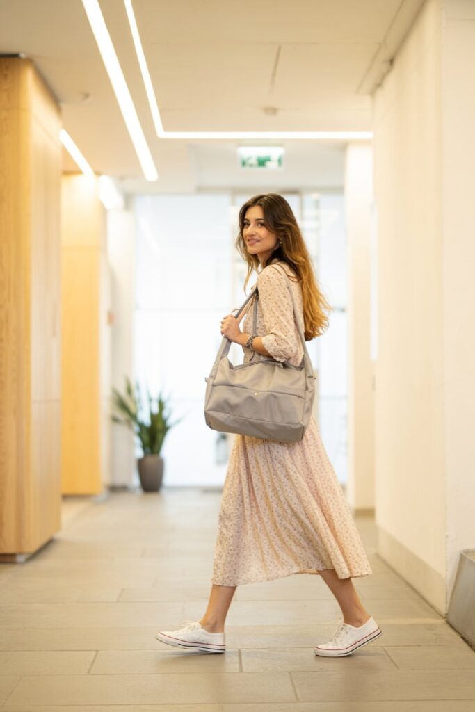 Trendy girl walking through a hallway in a soft floral midi dress and clean low-top sneakers, hair down, light office bag in hand