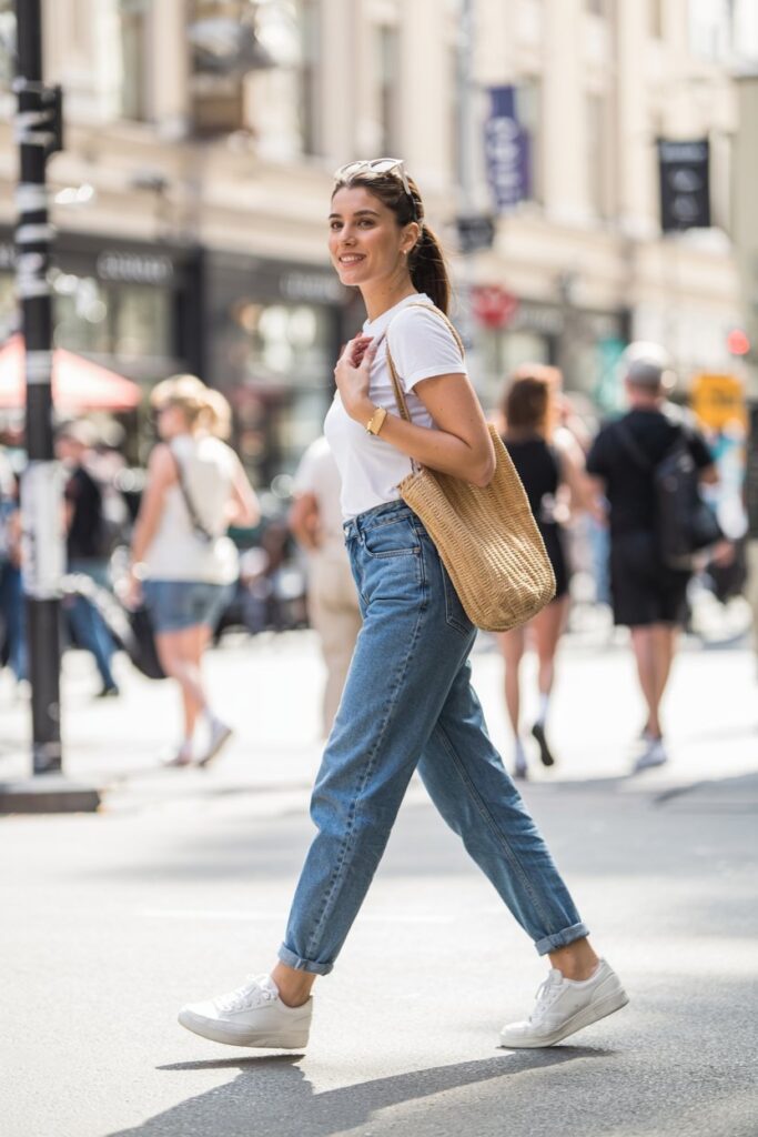 Woman in classic high-rise blue jeans and clean white tennis shoes, paired with a tucked-in white T-shirt, walking confidently down a sunlit street