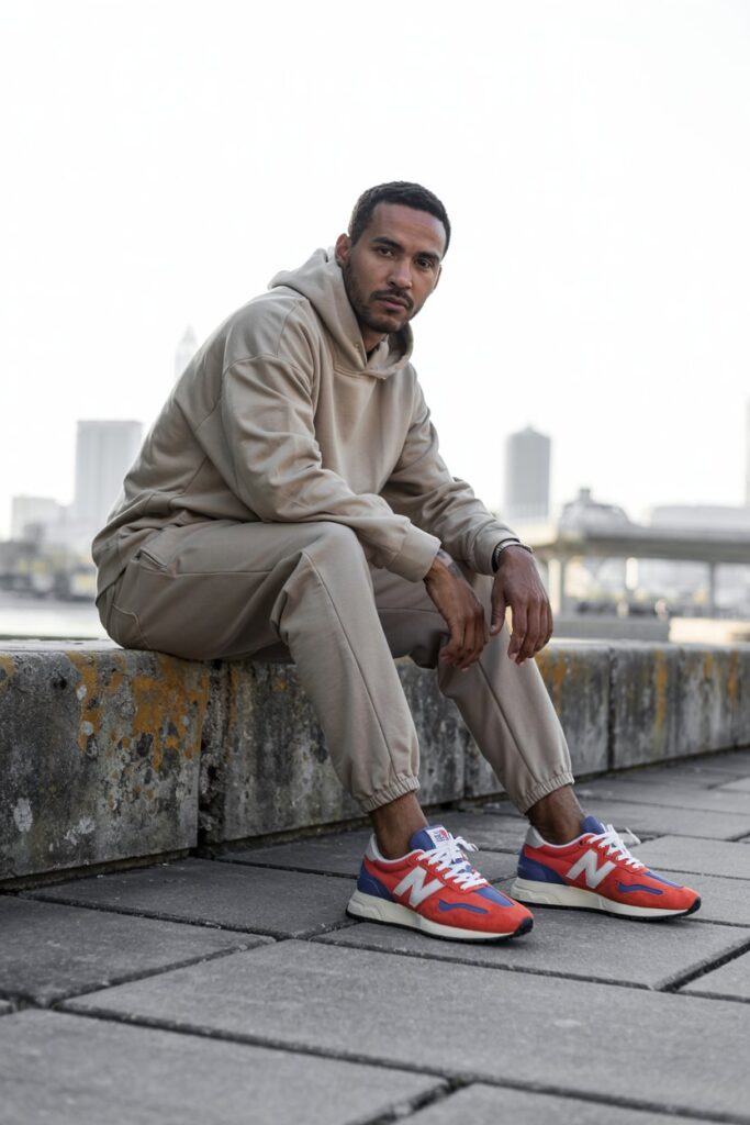 A stylish male model sitting on a low concrete ledge, beige joggers and hoodie combo, with bold orange or blue NB 327s clearly in focus