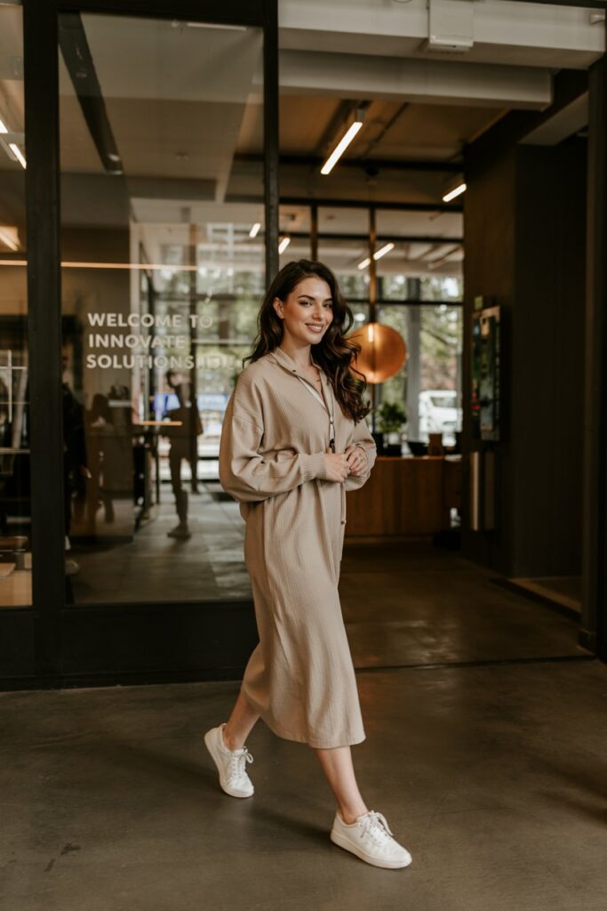 Trendy girl in a beige midi sweater dress with sleek leather sneakers, walking through a modern office lobby