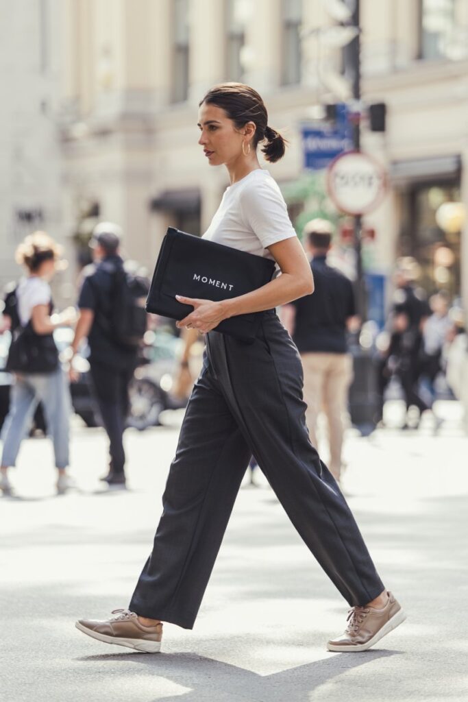 Trendy woman striding confidently in wide-leg trousers, tucked-in fitted tee, and sleek leather sneakers, holding a laptop sleeve