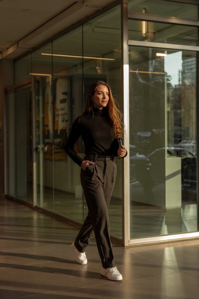 Trendy woman in high-waisted trousers, fitted black turtleneck, and platform white tennis shoes, confidently walking in a glass-walled hallway