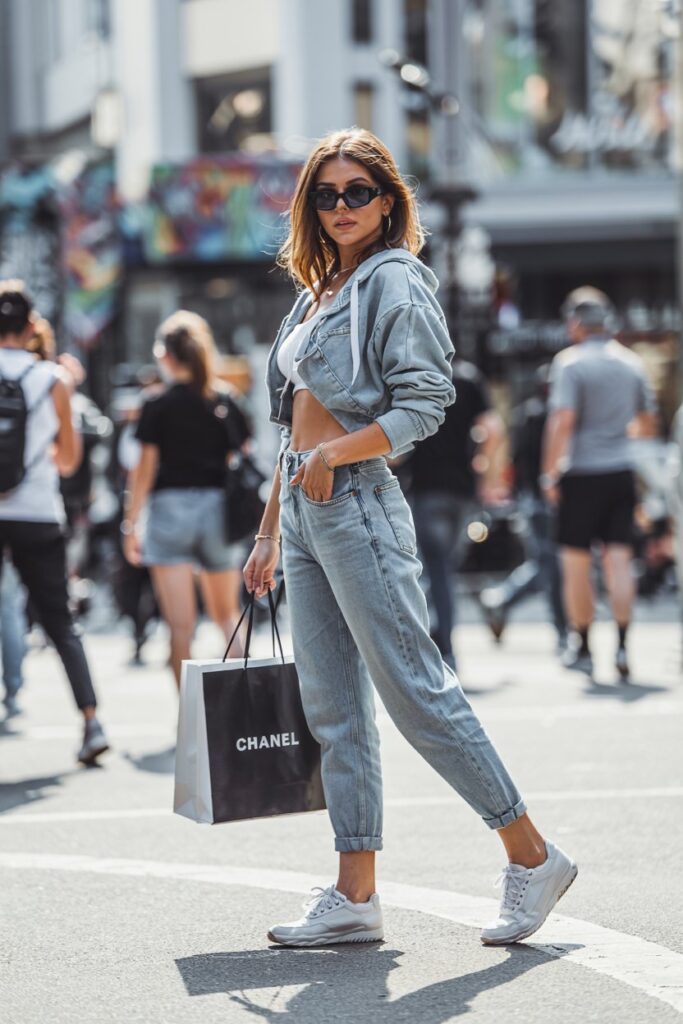 Trendy girl in light-wash mom jeans and retro-style white tennis shoes, cropped hoodie, sunglasses, posing on an urban sidewalk