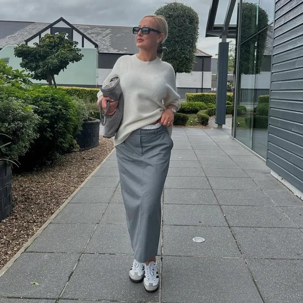 This outfit blends elegance with street edge. A soft white knit sweater pairs seamlessly with a tailored grey maxi skirt, while white Adidas Sambas add that sporty-cool contrast.