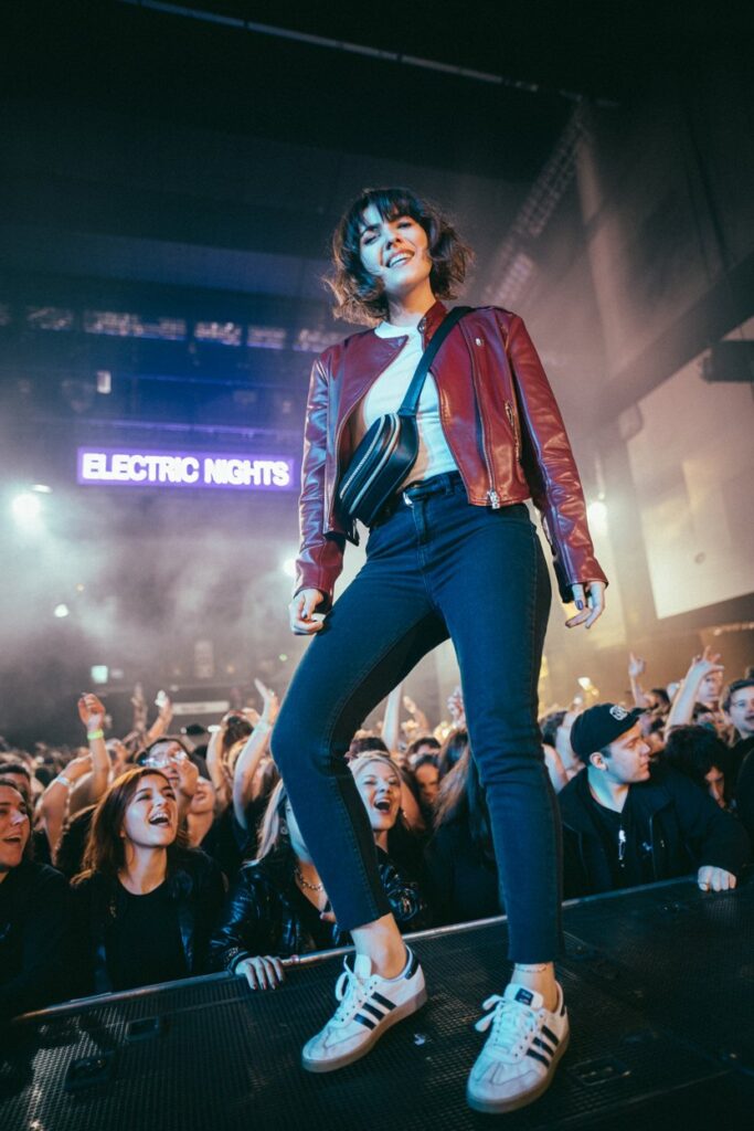 Edgy woman at a concert in burgundy leather jacket, black jeans, crossbody bag, and white Adidas Sambas	