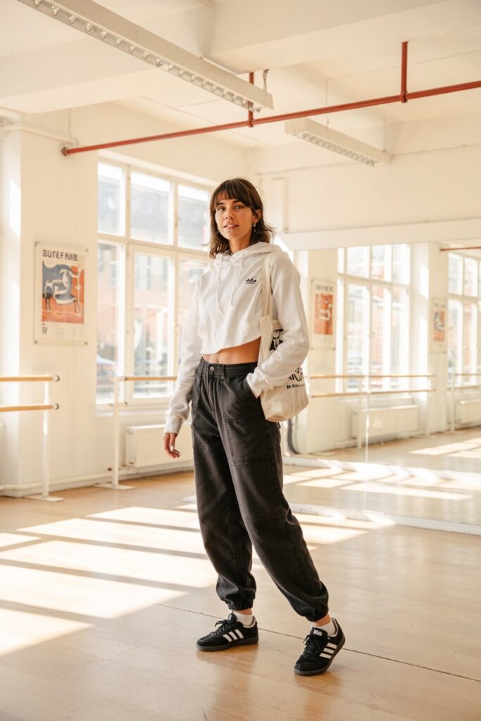 woman in black Adidas Samba sneakers, dark wash denim joggers, white cropped hoodie, white tote bag, colorful dance studio, nostalgic 90s-inspired workout fashion