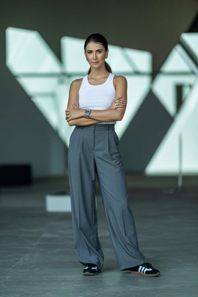 Bold woman at a design firm wearing gray wide-leg trousers, white tank, statement watch, and black Adidas Sambas	
