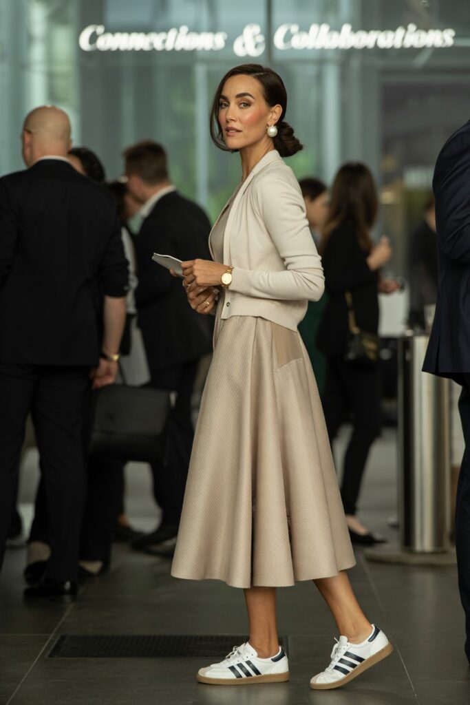 Elegant woman at a networking event wearing a beige midi dress, fitted cardigan, pearl earrings, and white Adidas Sambas	