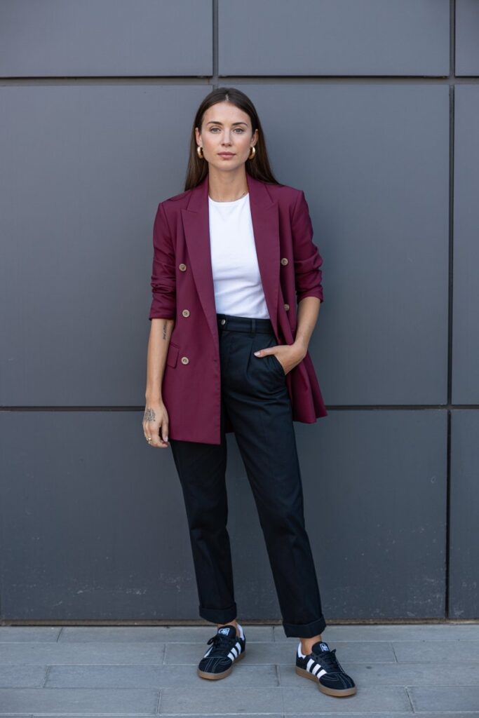 Confident woman at a corporate event in burgundy blazer, white tee, black trousers, gold stud earrings, and black Adidas Sambas	