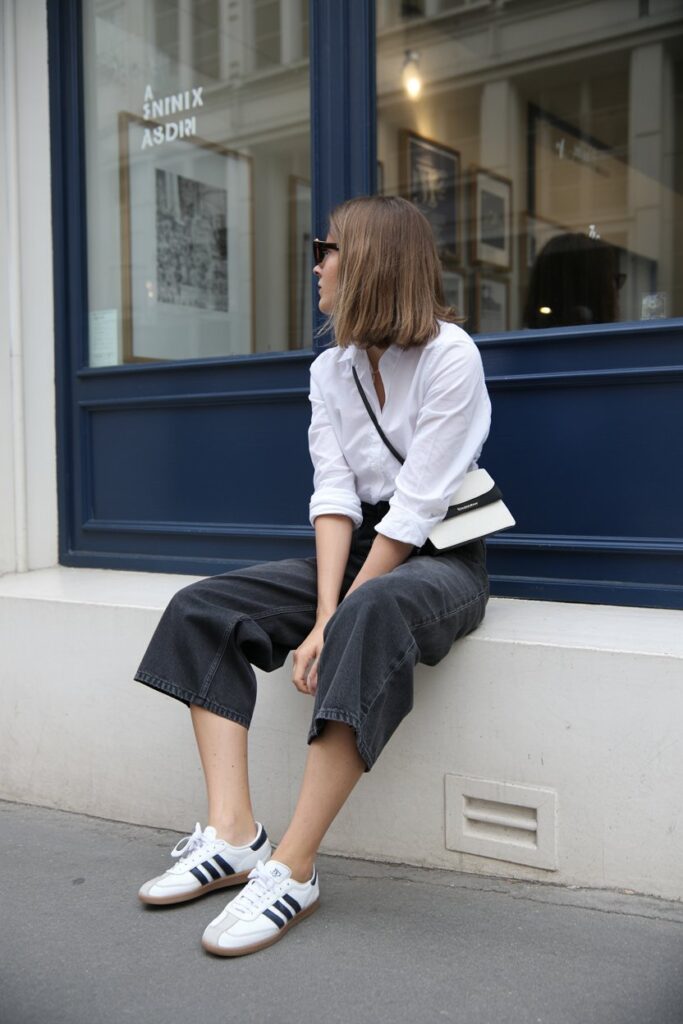 Parisian-style woman sitting outside a gallery in high-waisted oversized black denim shorts, white blouse, crossbody bag, and white Adidas Sambas