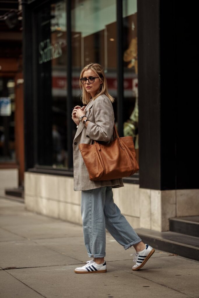 Casual downtown style: woman in oversized gray blazer, light blue jeans, Adidas Sambas, leather tote bag, walking on a city sidewalk