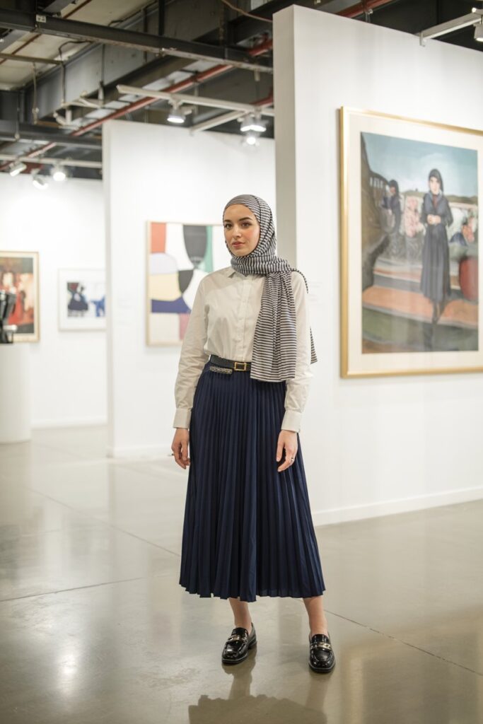 Preppy hijab look: navy pleated midi skirt, white button‑down shirt, striped jersey hijab, black loafers, art gallery or panel event setting