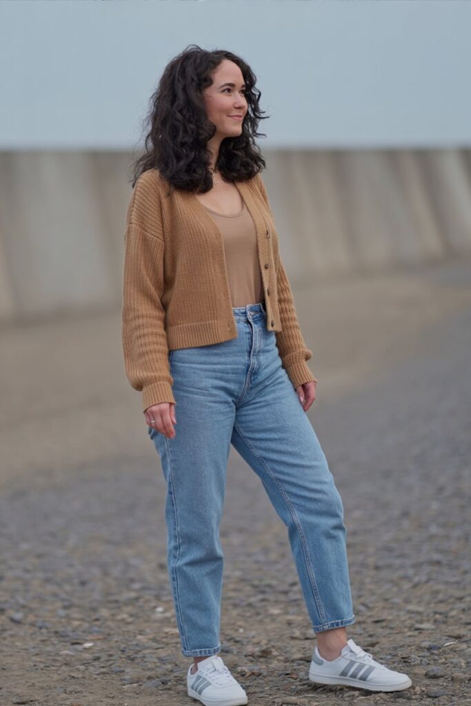 petite woman standing casually, tan cropped cardigan, high-waist light blue jeans, white Adidas Sambas visible, everyday outfit realism.