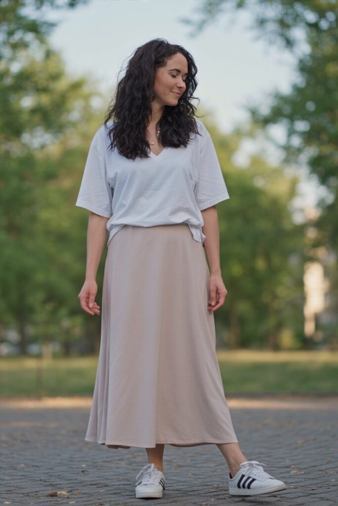 woman in white graphic tee tucked into flowy beige maxi skirt, white Adidas Sambas visible, relaxed full-body style.