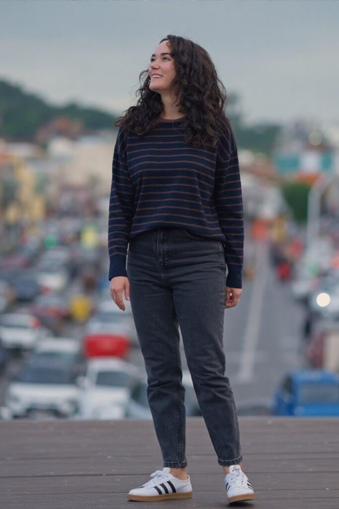 woman in striped navy sweater and black jeans, white Adidas Sambas visible, classic casual outfit.