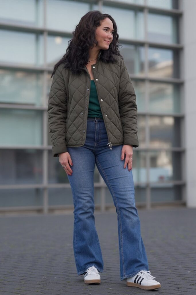 woman in olive quilted jacket and medium-wash flared jeans, white Adidas Sambas visible, standing naturally.