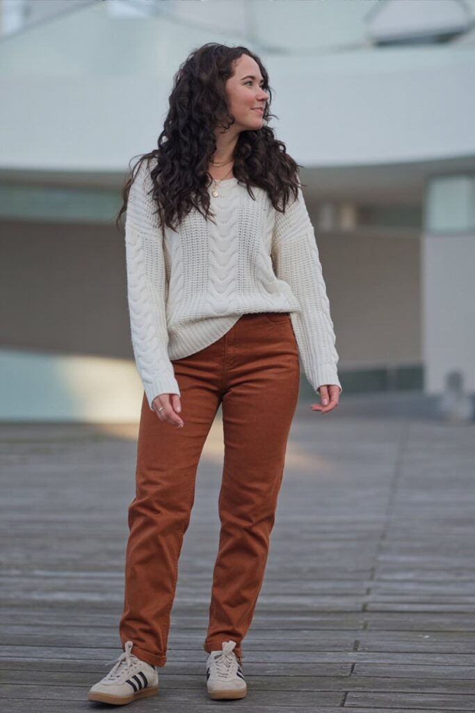 woman in ivory cable-knit sweater and rust corduroy pants, beige Adidas Sambas visible, standing naturally, full-body proportion.