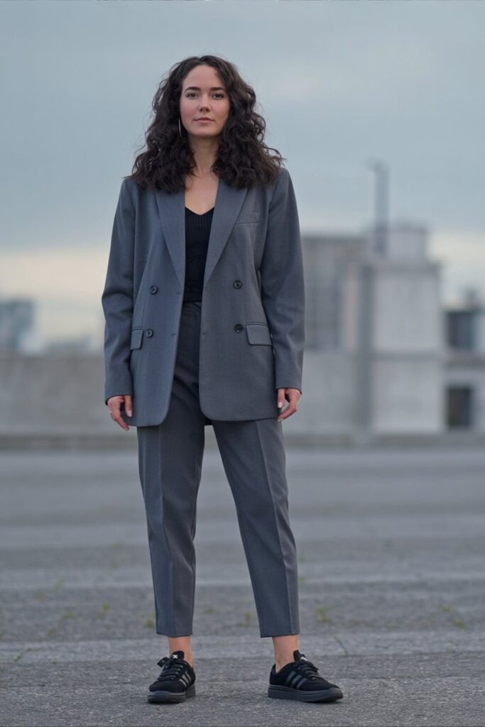 woman in oversized gray blazer and ankle trousers with black Adidas Sambas, standing straight in minimalist urban scene.