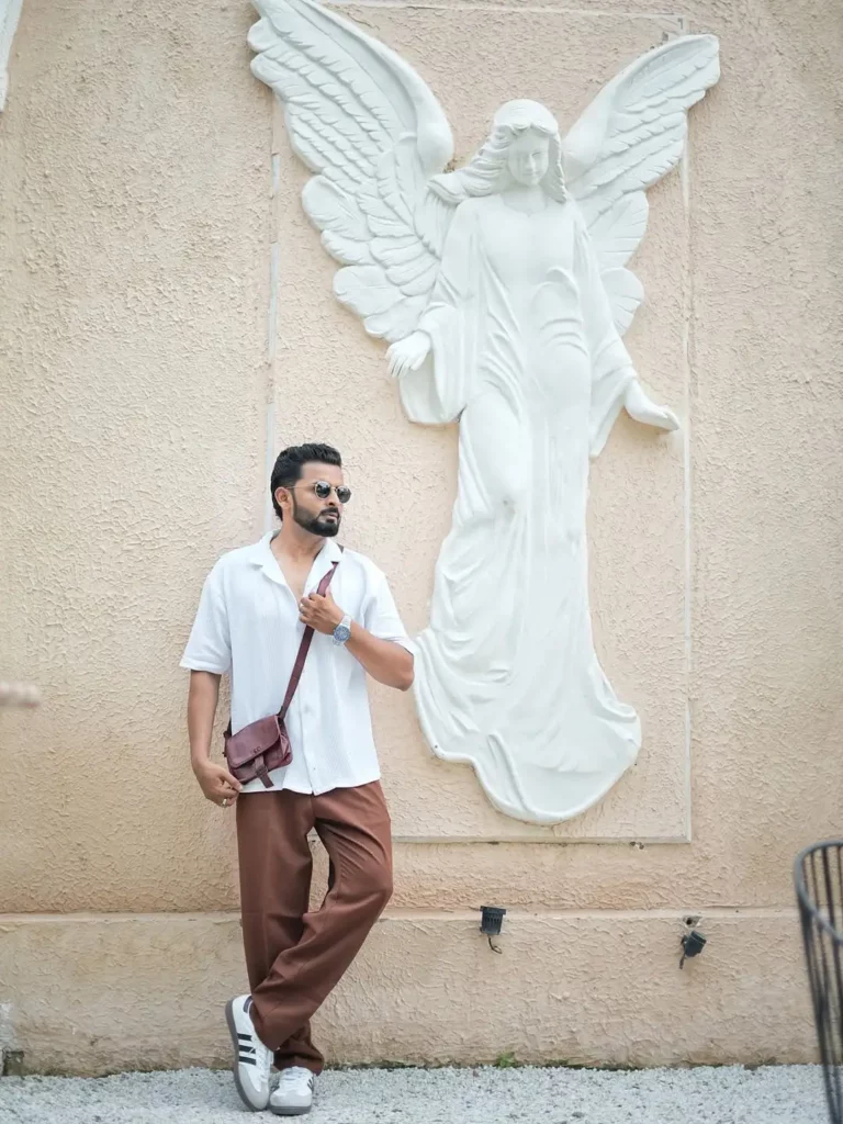 The white open-collar shirt and flowy brown trousers create a relaxed but elevated base. Paired with the white Adidas Sambas